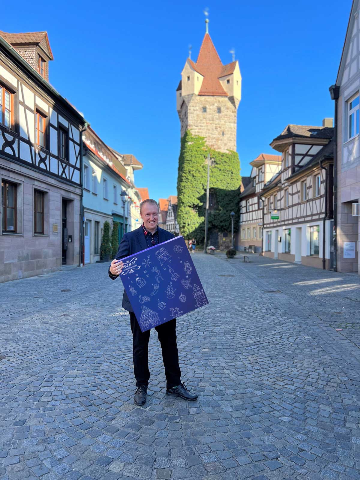 Christian Kindler steht vor dem Fehnturm in Herzogenaurach und hält den HEIMATadventskalender 2023 in der Hand.